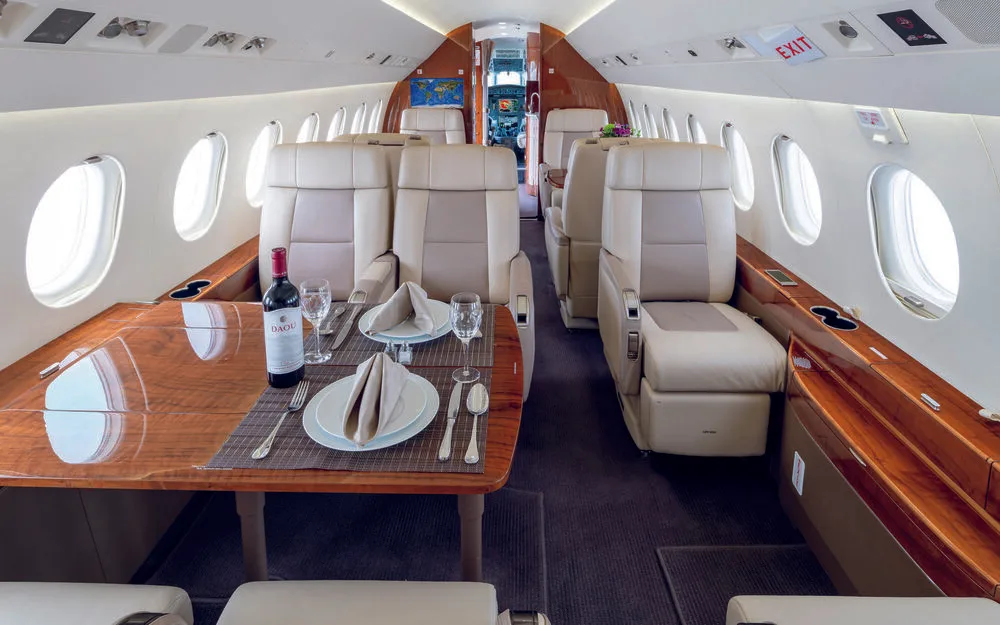 The luxurious cabin interior of a business jet, featuring beige leather seats, a polished wooden dining table set with wine and place settings, and a world map display.