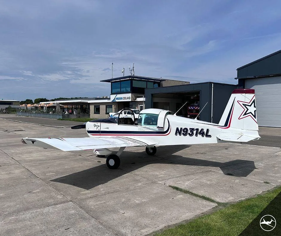 A white Grumman AA-5B Tiger aircraft with black and red stripes, registration N9314L, parked on concrete in front of an airport building.