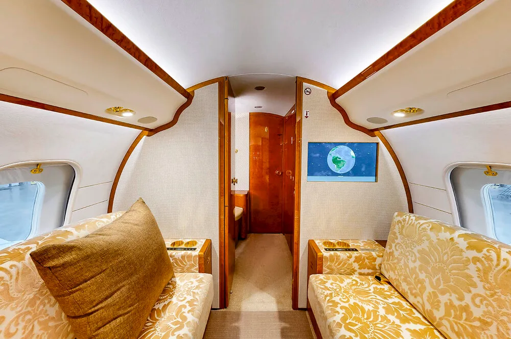 The aft section of the business jet's cabin, showing two patterned couches facing each other with a wall-mounted screen displaying a globe.
