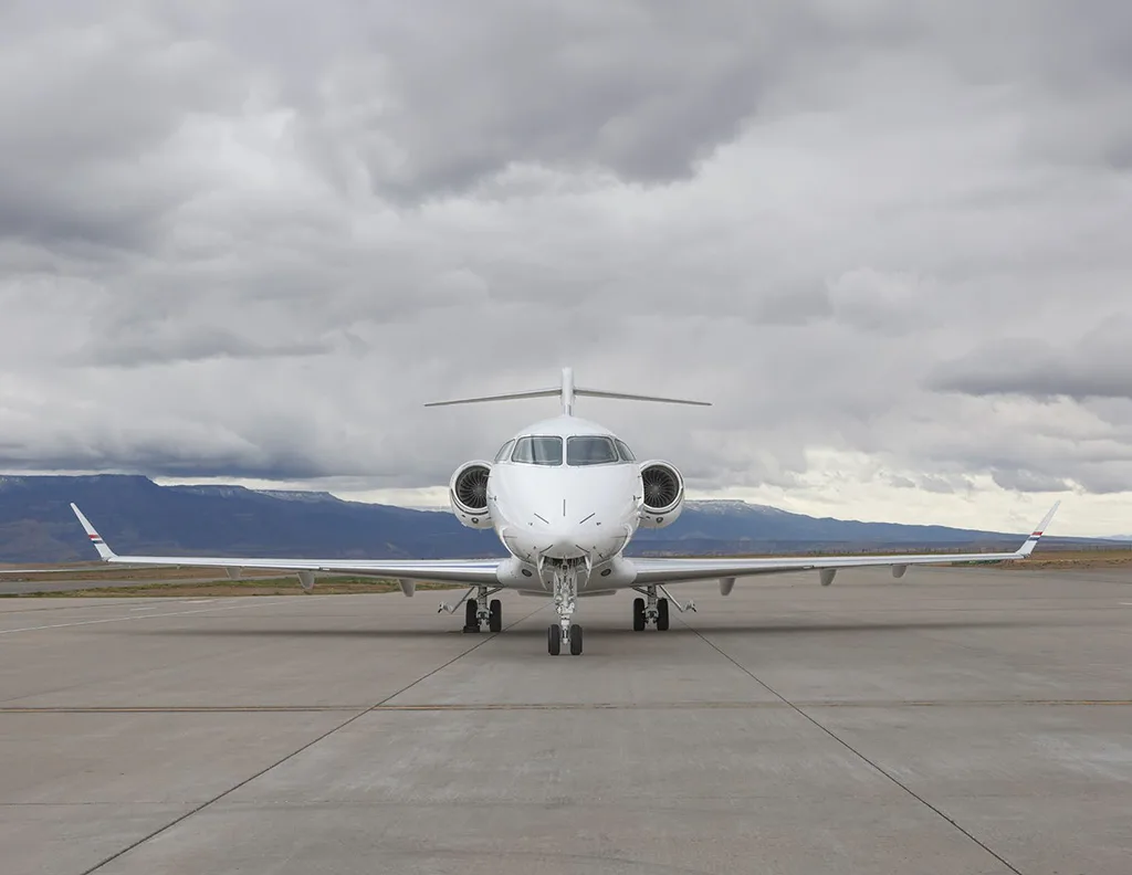 A head-on view of a white Bombardier Challenger 350 business jet parked on an airfield under a cloudy sky.