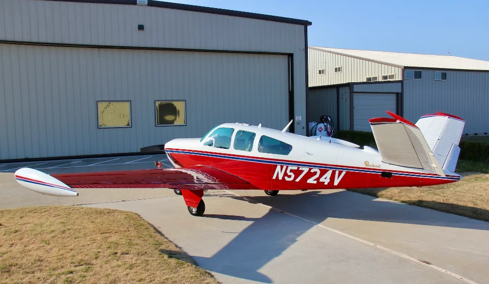 A red and white Beechcraft Bonanza N5724V parked on an airfield next to a hangar.