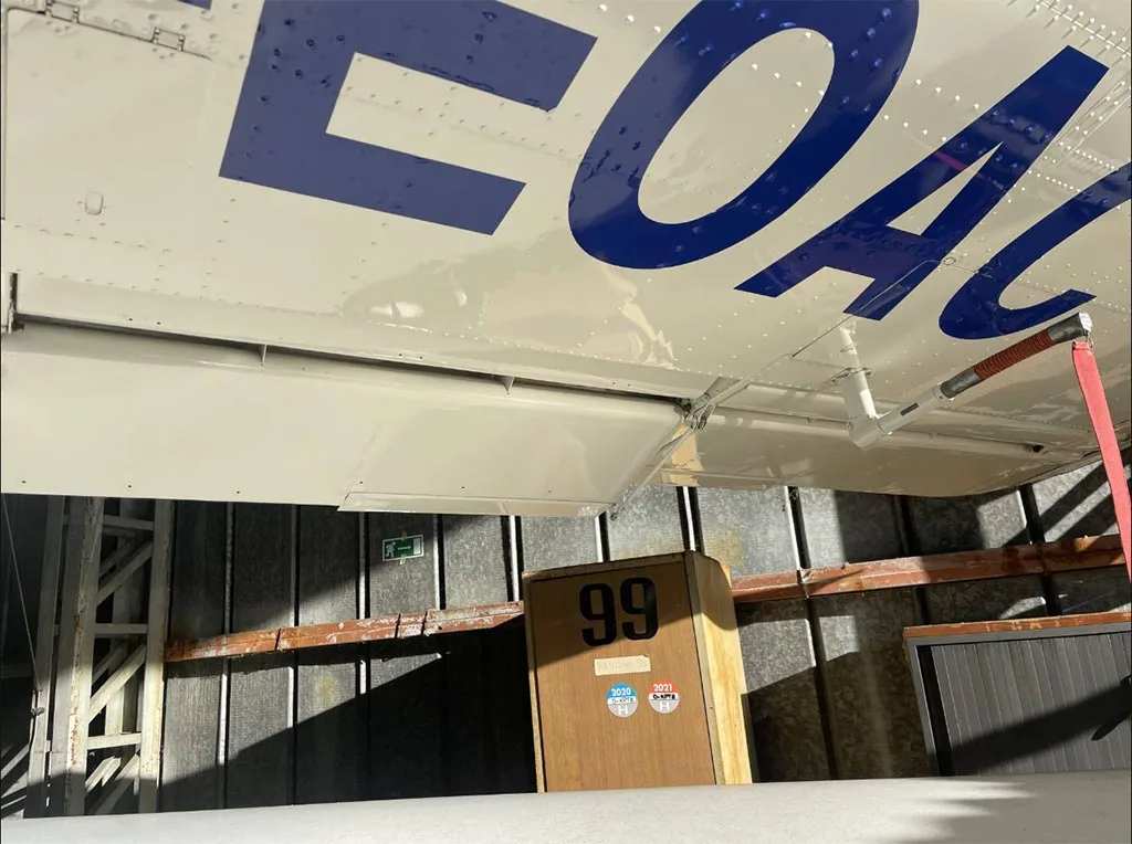 The underside of the wing of a white aircraft, D-EOAC, showing flaps and part of the fuselage, inside a hangar.