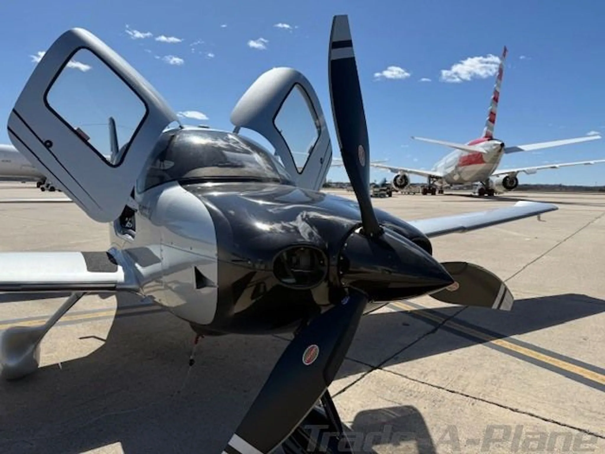 A black and silver Cirrus SR22 GTS Turbo aircraft with open doors is parked on a tarmac with a commercial airliner in the background.