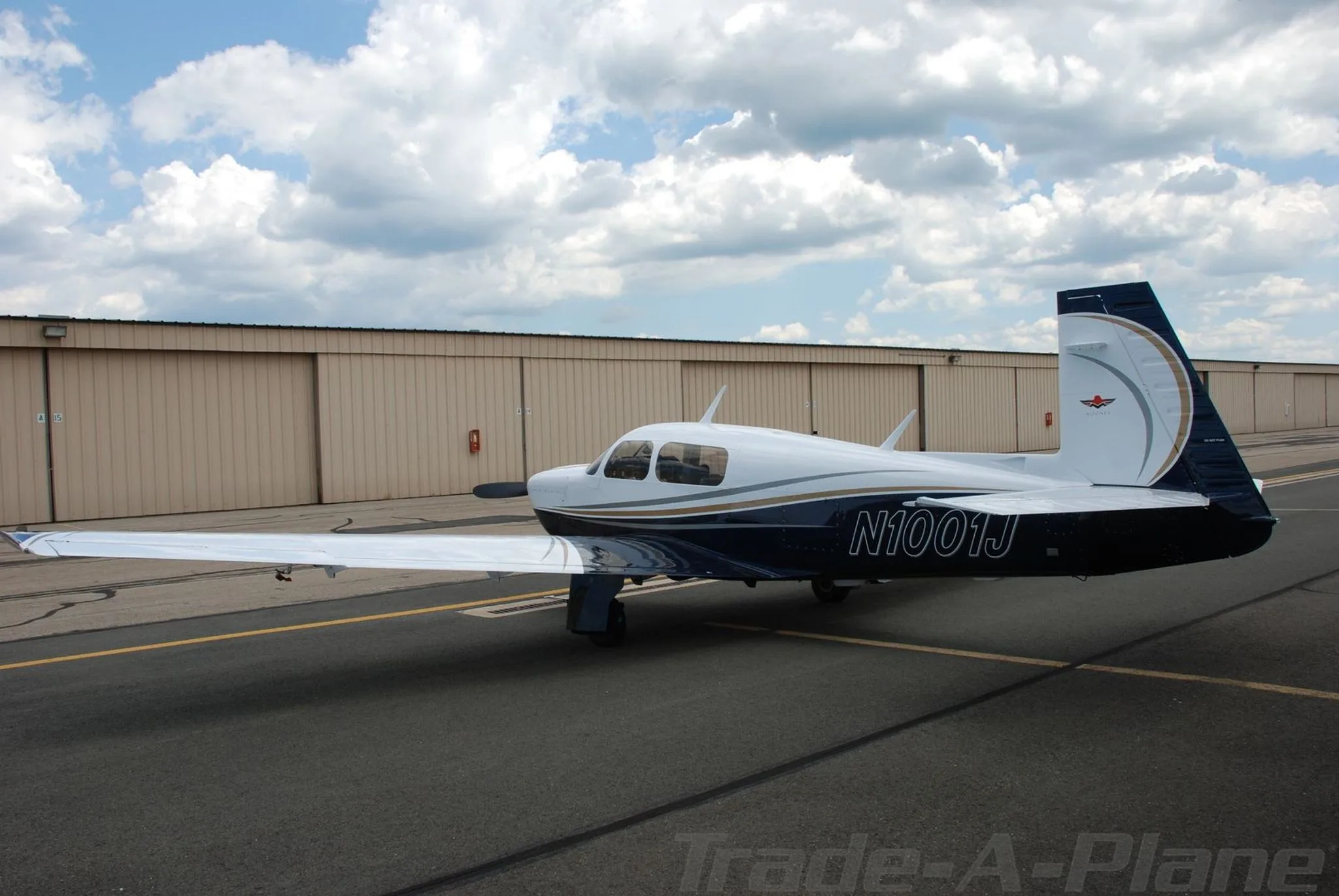 A three-quarter rear view of the white and blue Mooney aircraft (N1001J) parked on a tarmac.