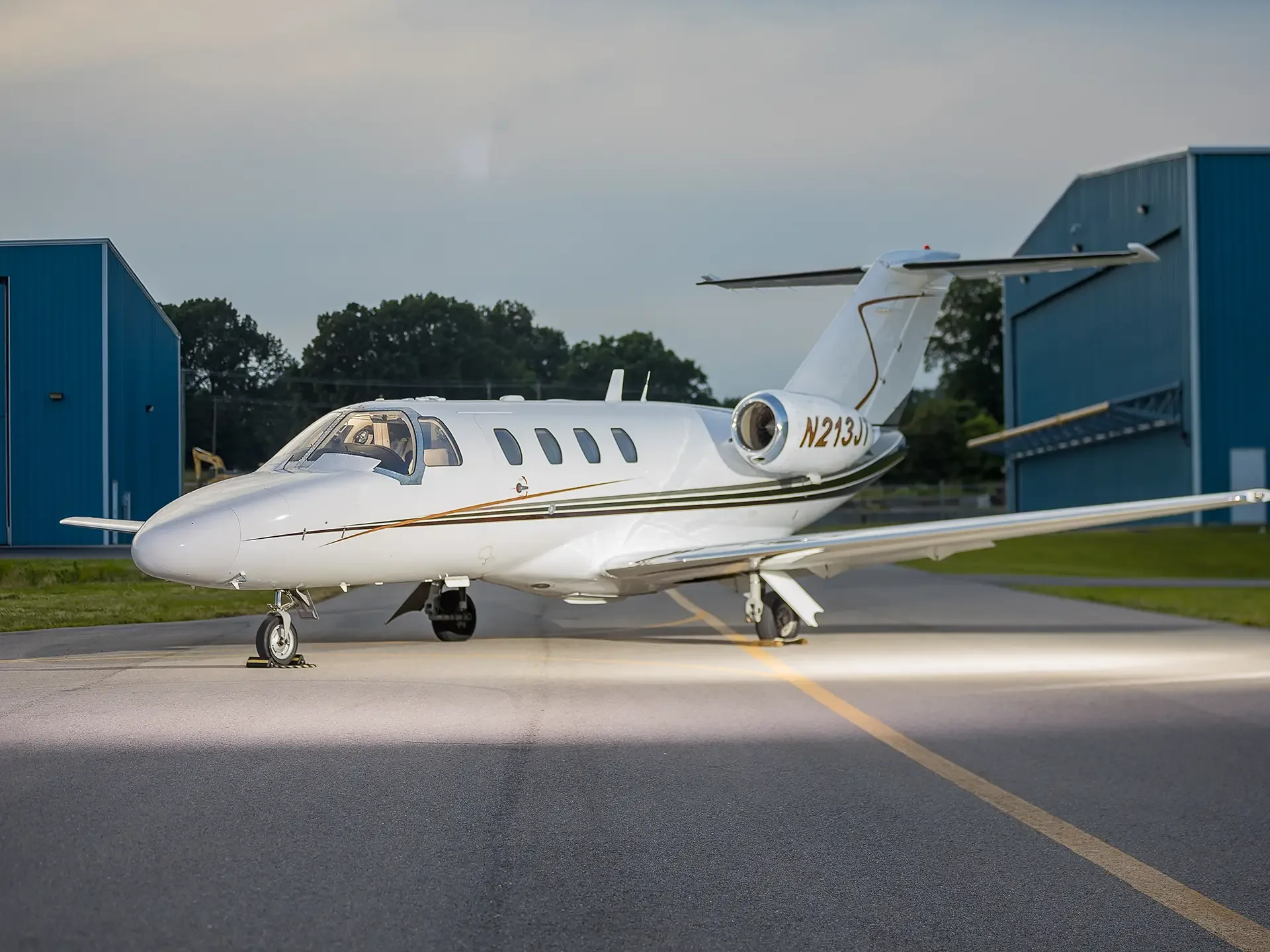 A white Cessna Citation business jet with gold and green stripes is parked on a tarmac at dusk, with hangars in the background.