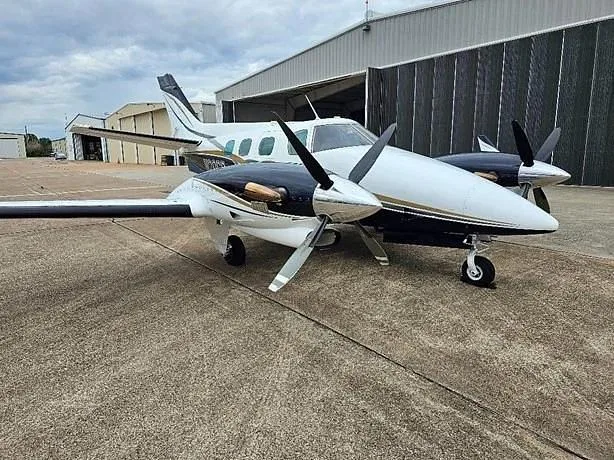 A white and black twin-engine propeller aircraft (N20CG) parked on an airfield next to a hangar.