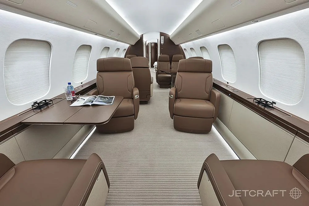 A view down the aisle of a business jet cabin shows brown leather seats and tables set with water and a magazine.
