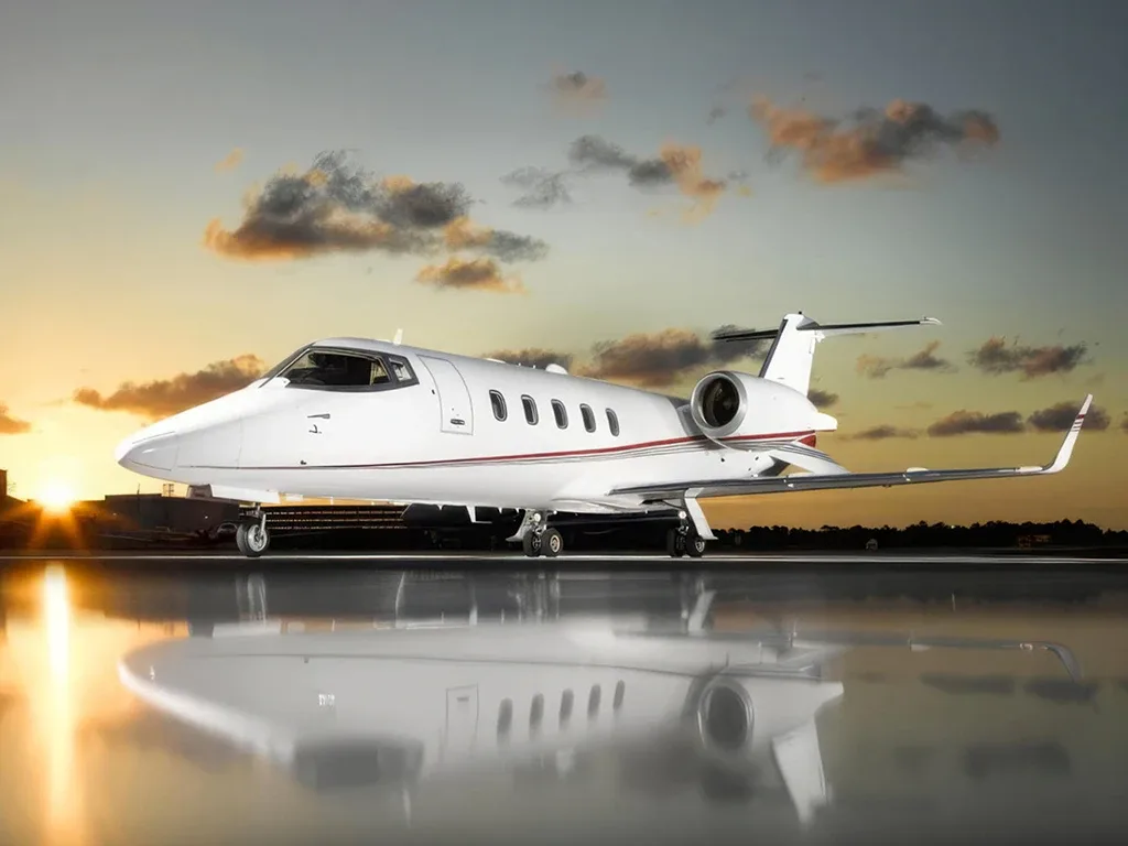 A white business jet with red stripes is parked on a reflective surface at sunset, with dramatic clouds in the sky.
