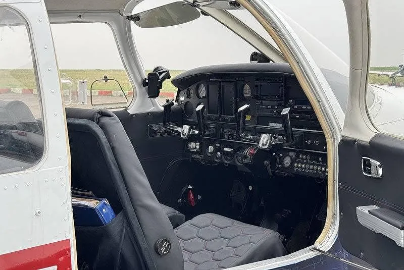 An angled view into the cockpit of a Piper PA-28 aircraft with the door open, showing the instrument panel and pilot's seat.