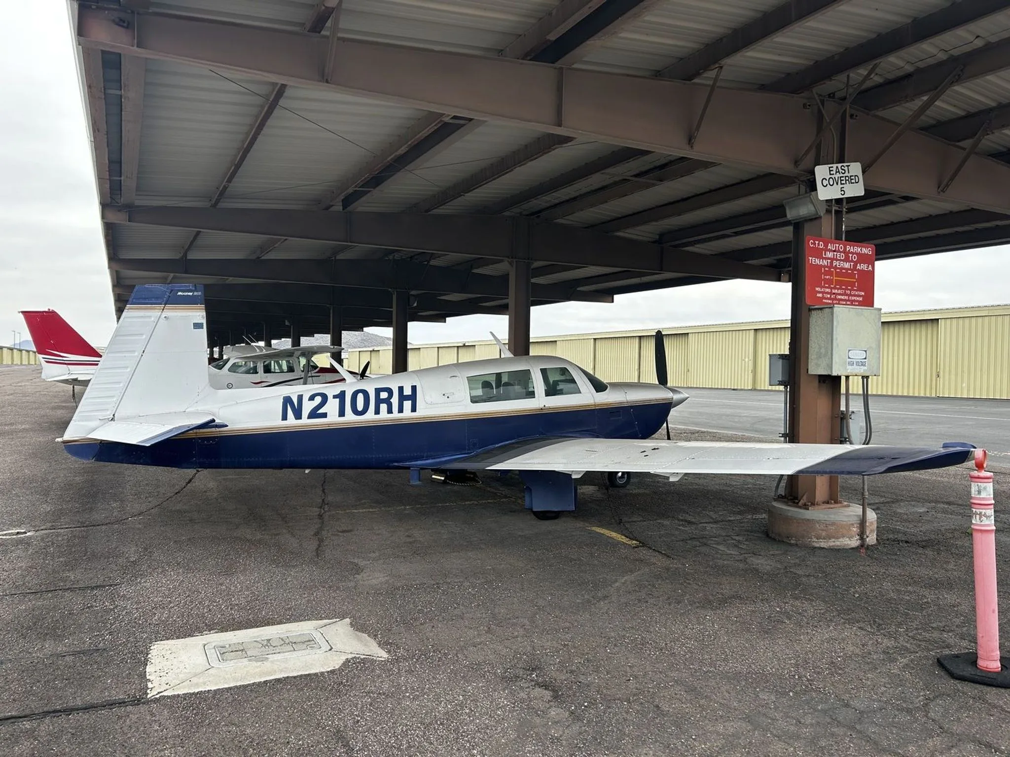A blue and white single-engine aircraft, N210RH, parked under a covered shelter at an airport.