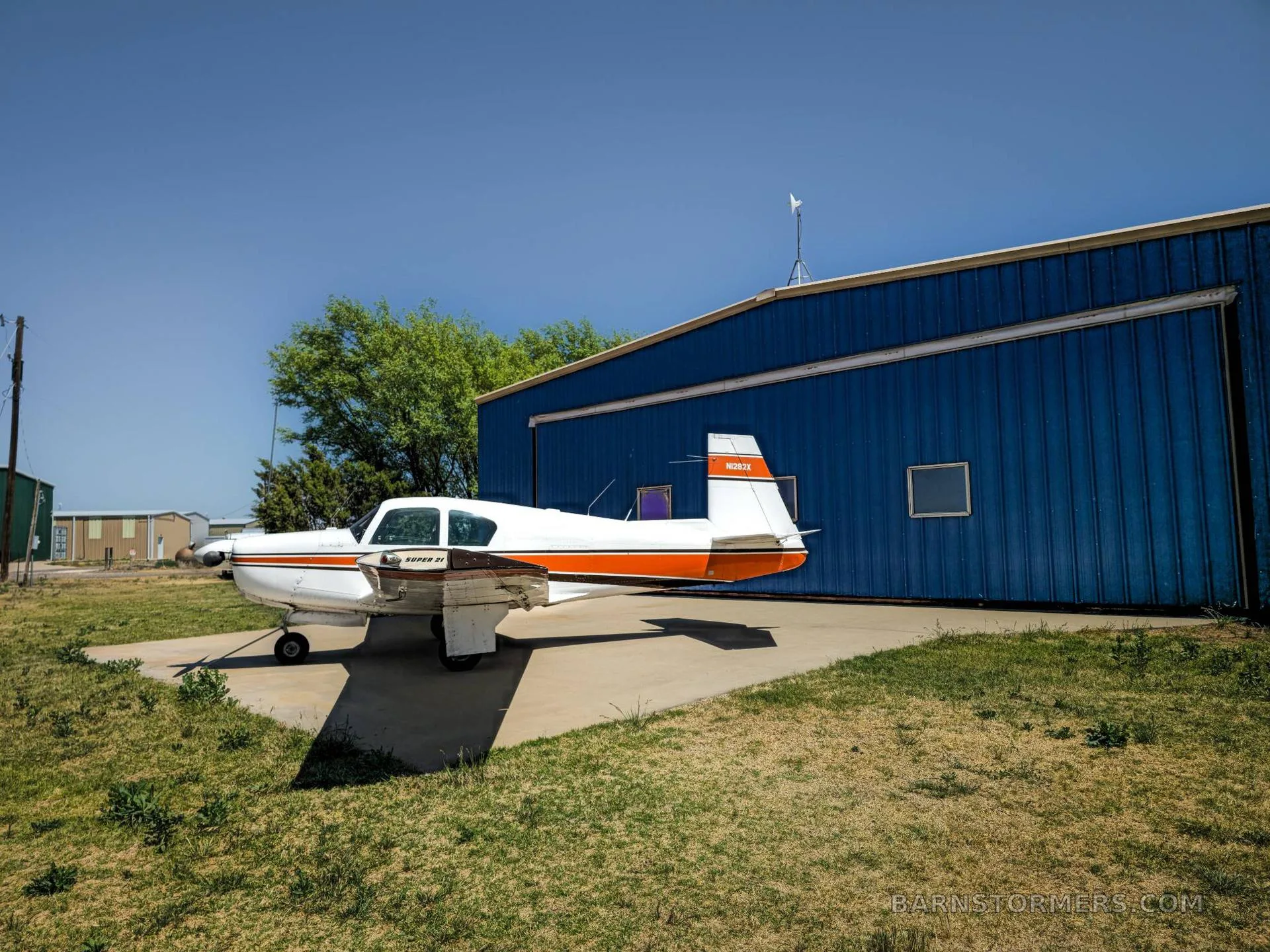 A white and orange single-engine aircraft (N282X) with "SUPER 21" text on its side, parked on a concrete pad next to a blue hangar.