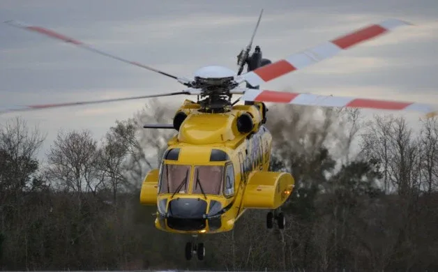 A yellow search and rescue helicopter with red and white striped rotor blades hovers above a treeline.