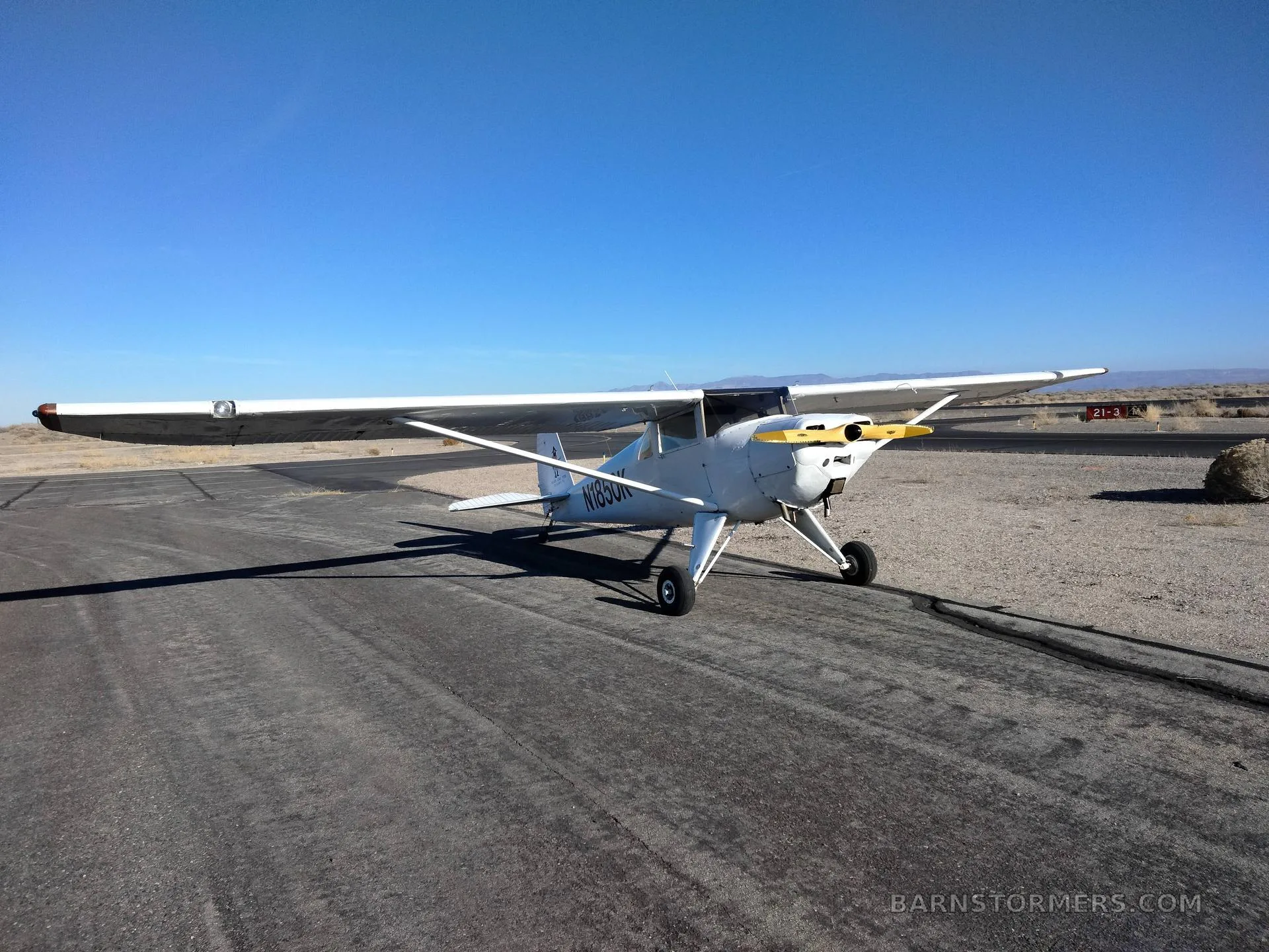 A white Luscombe 8A aircraft with a yellow propeller is parked on an asphalt runway in a desert setting.