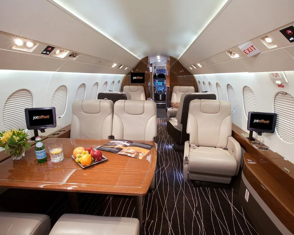 A view inside the business jet cabin, showing beige leather seating, a wooden table set with fruit and drinks, and an "EXIT" sign above the door.