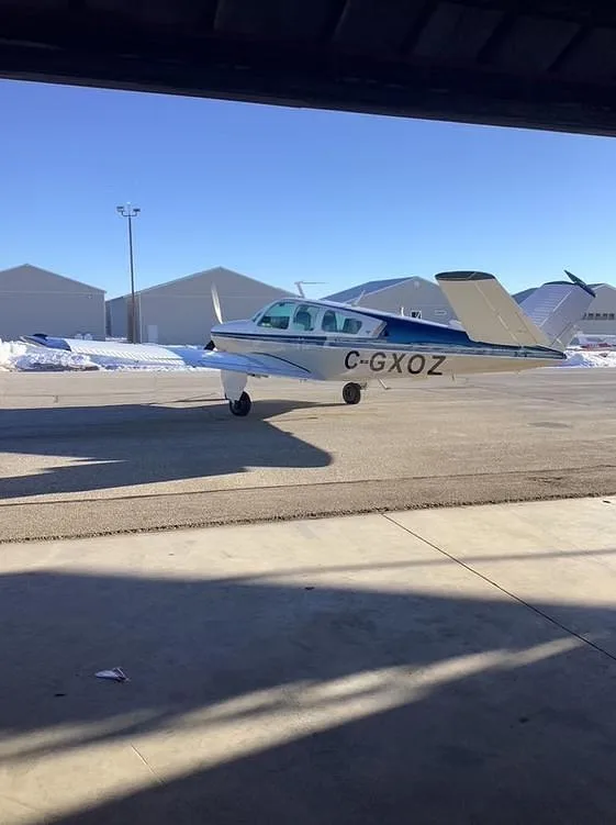 A beige and blue single-engine aircraft with registration C-GXOZ is parked on an airfield, viewed from inside a hangar.