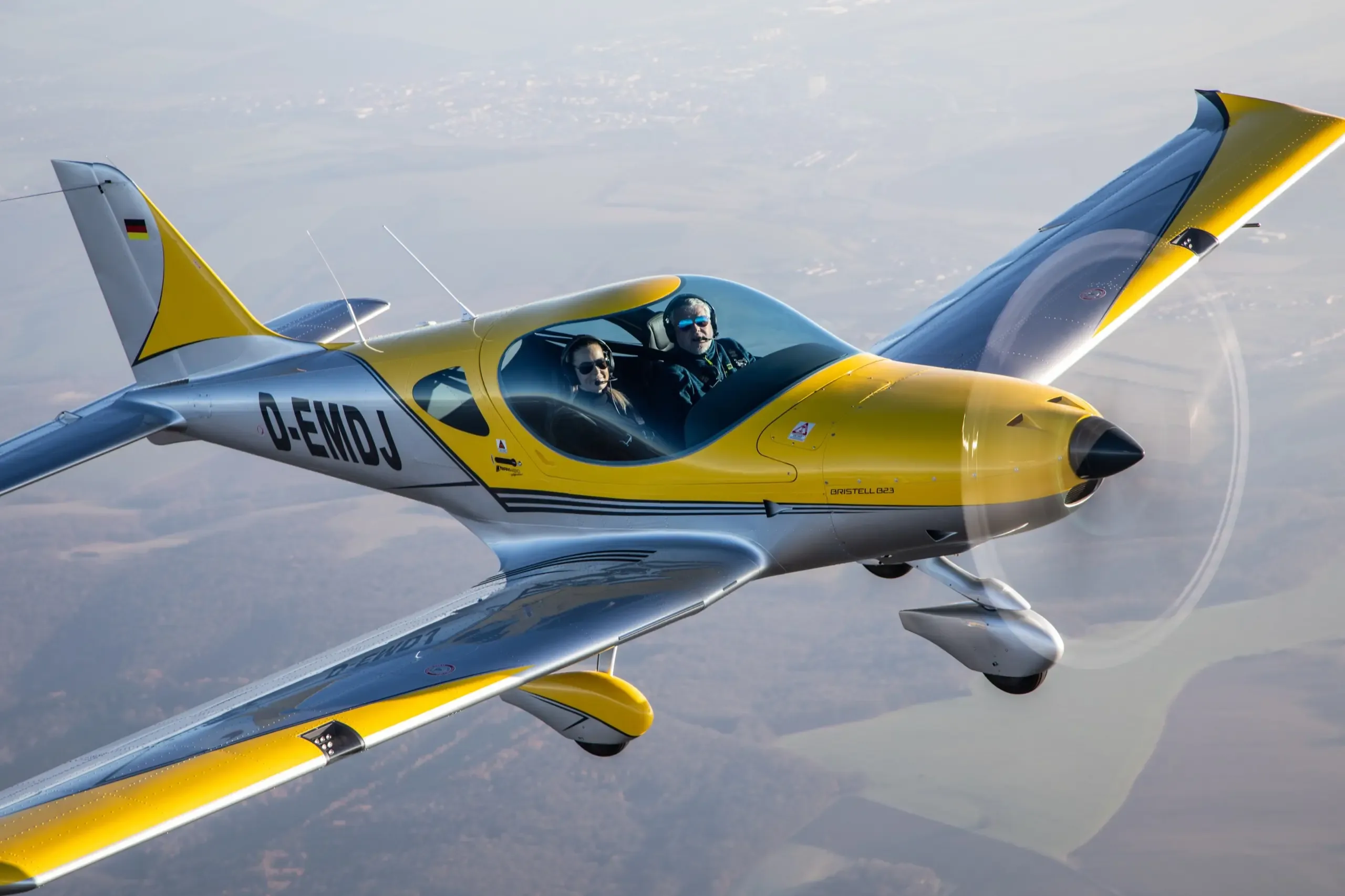 An aerial view of a yellow and silver Bristell B23 aircraft with two occupants in flight over a hazy landscape.