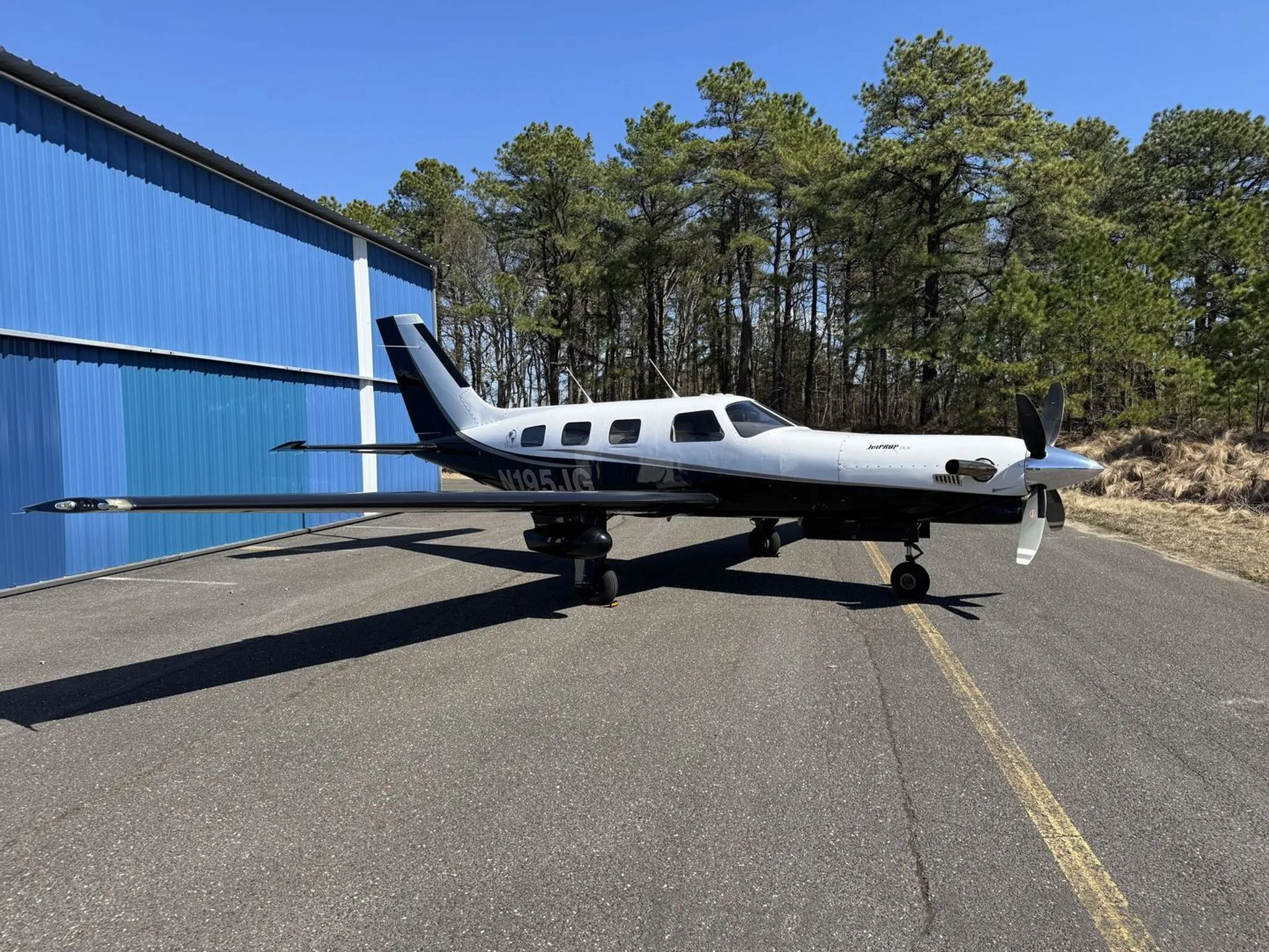 A white and black Piper M-class aircraft, tail number N195JG, parked on an asphalt ramp next to a blue hangar.