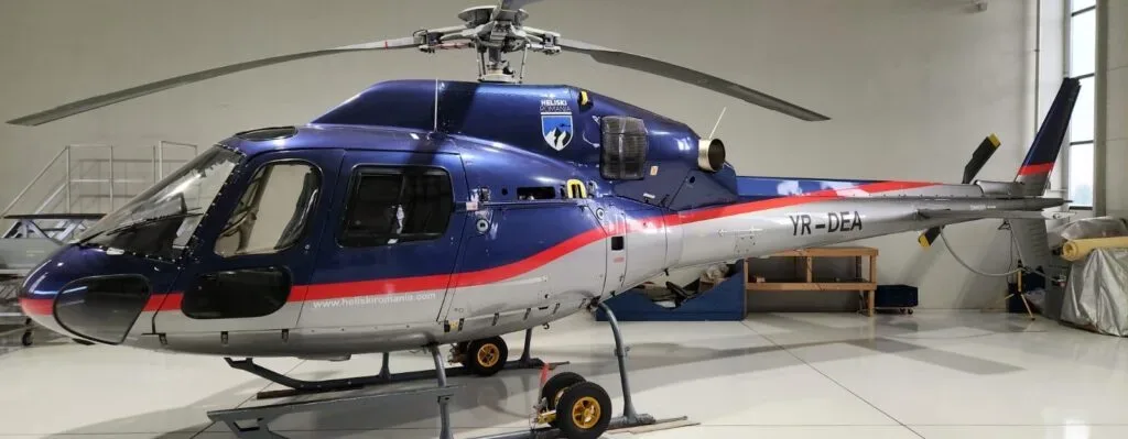 A blue and silver Airbus H125 helicopter with a red stripe and 'YR-DEA' registration, parked inside a hangar.