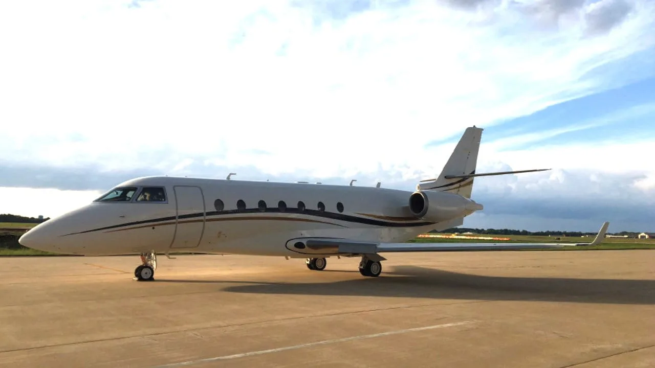 A white business jet with dark and gold stripes parked on an airfield at sunset.