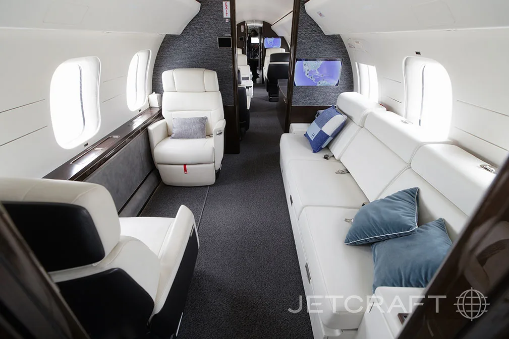 The luxurious interior of a private jet cabin with white leather seating, a couch, and blue accent pillows.