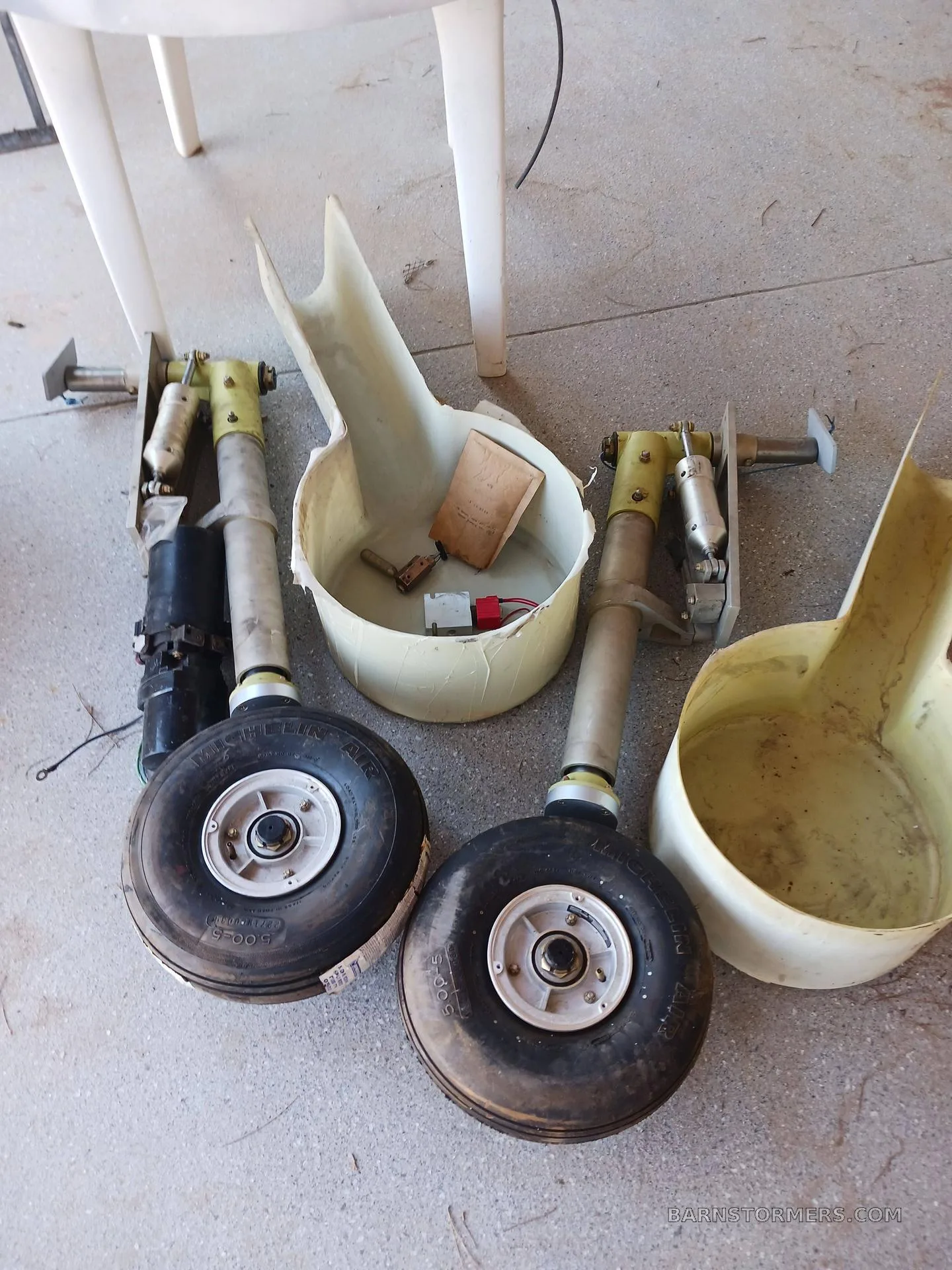 Two aircraft main landing gear struts with wheels and associated fairings are shown on a concrete floor.
