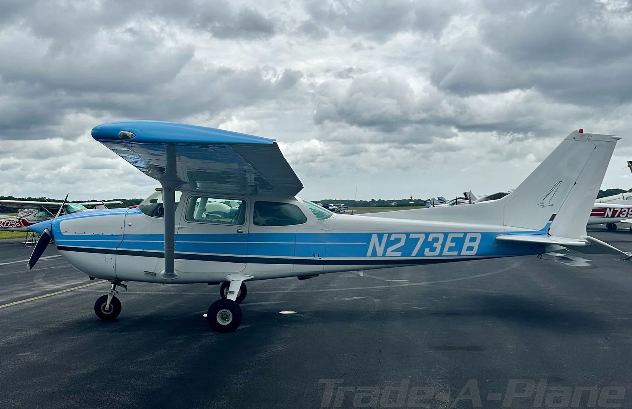 A white and blue Cessna 172 aircraft, registration N273EB, parked on an asphalt ramp under a cloudy sky.