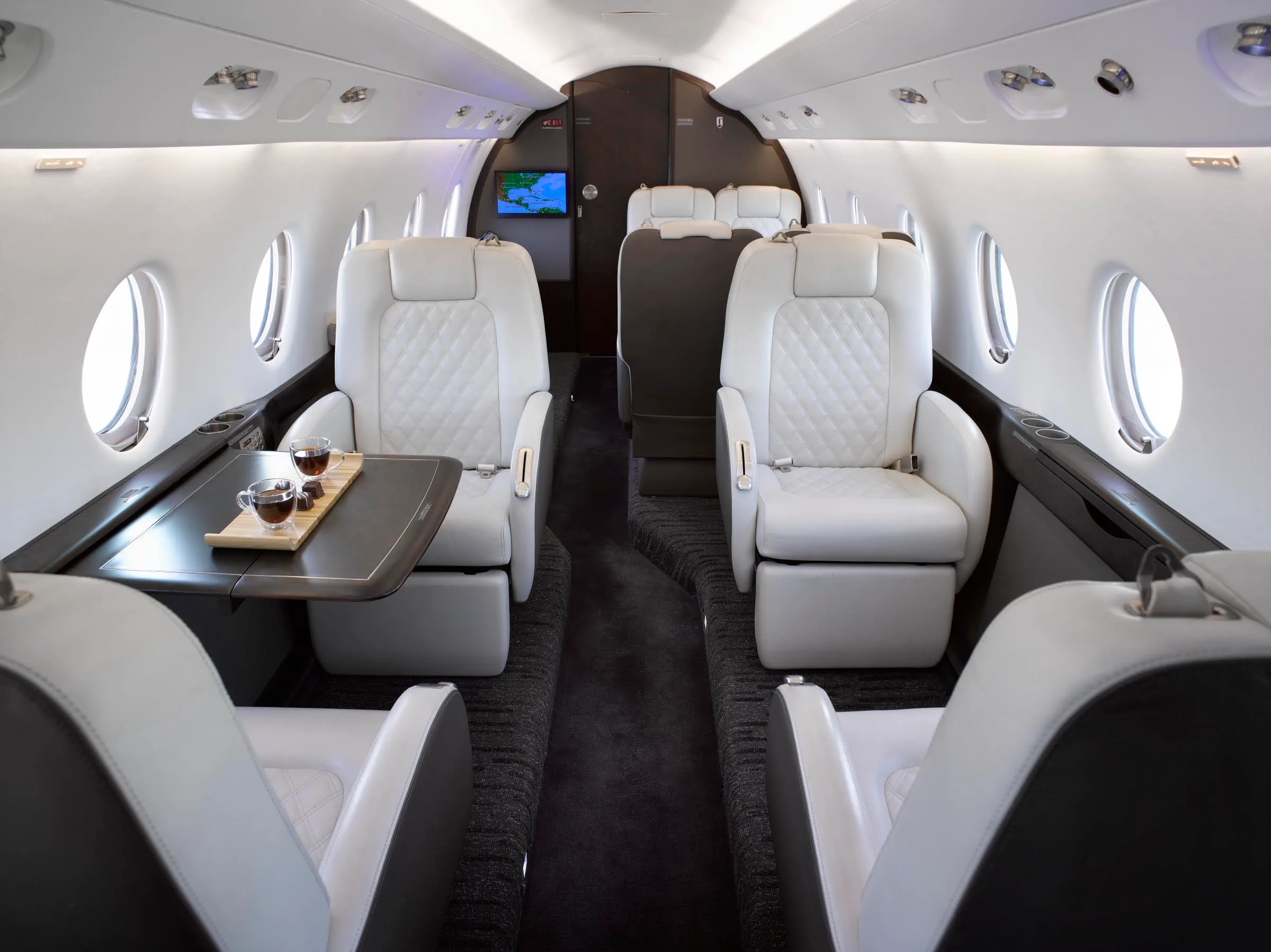 The elegant interior of a private jet cabin features rows of white quilted leather seats and a table with two cups of coffee.