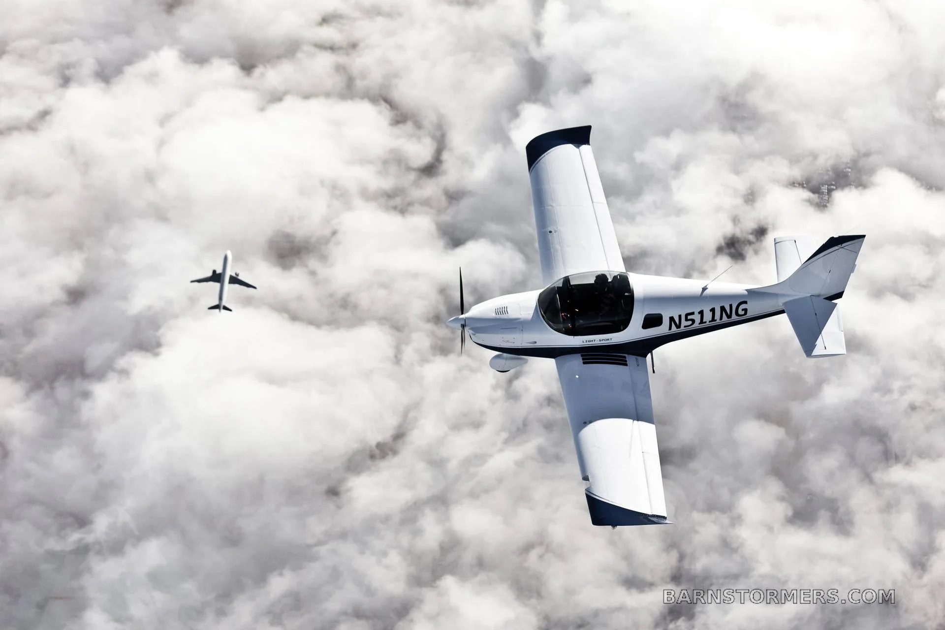 A white Sling 2 aircraft (N511NG) with black accents flies above clouds, with another aircraft in the distance.