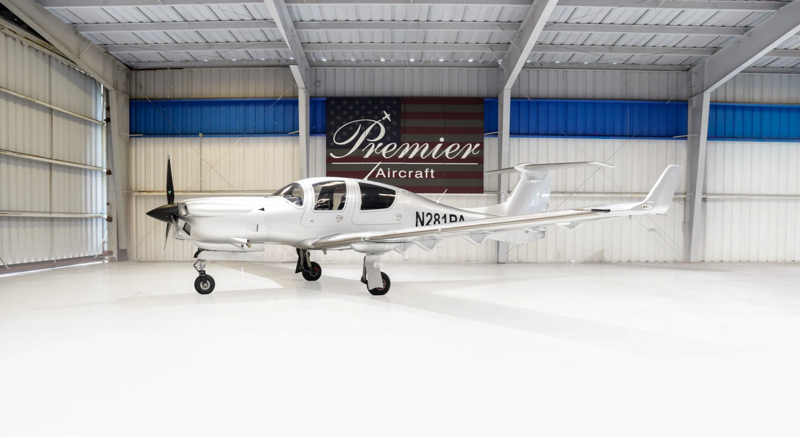 A silver Diamond DA42 Twin Star aircraft, registration N281PA, is parked inside a hangar with a 'Premier Aircraft' banner in the background.