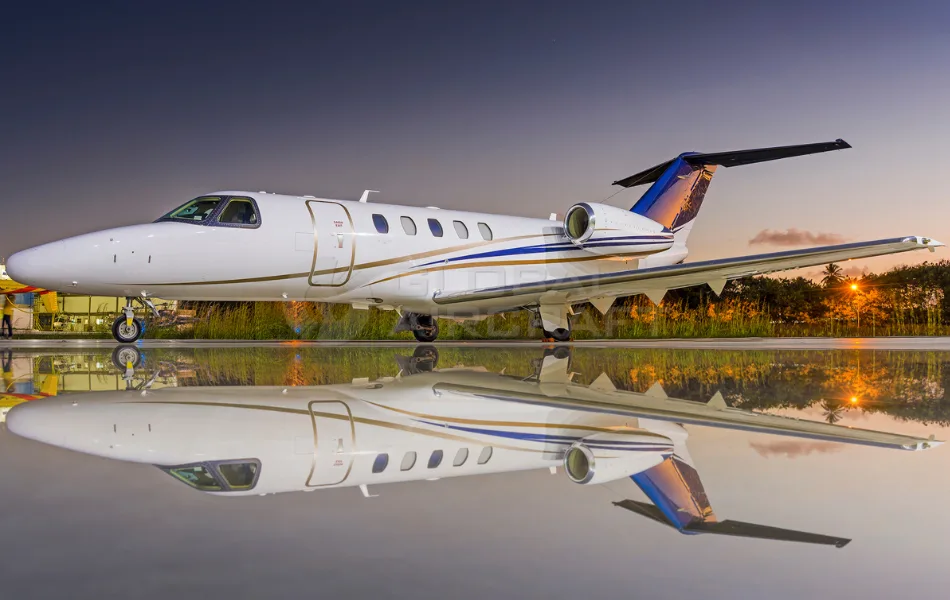A white business jet with gold and blue stripes and a colorful tail, parked on a reflective surface at dusk.