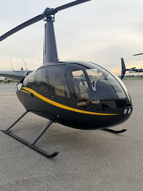 A black Robinson R44 helicopter with a yellow stripe, viewed from the front-right on an airfield.