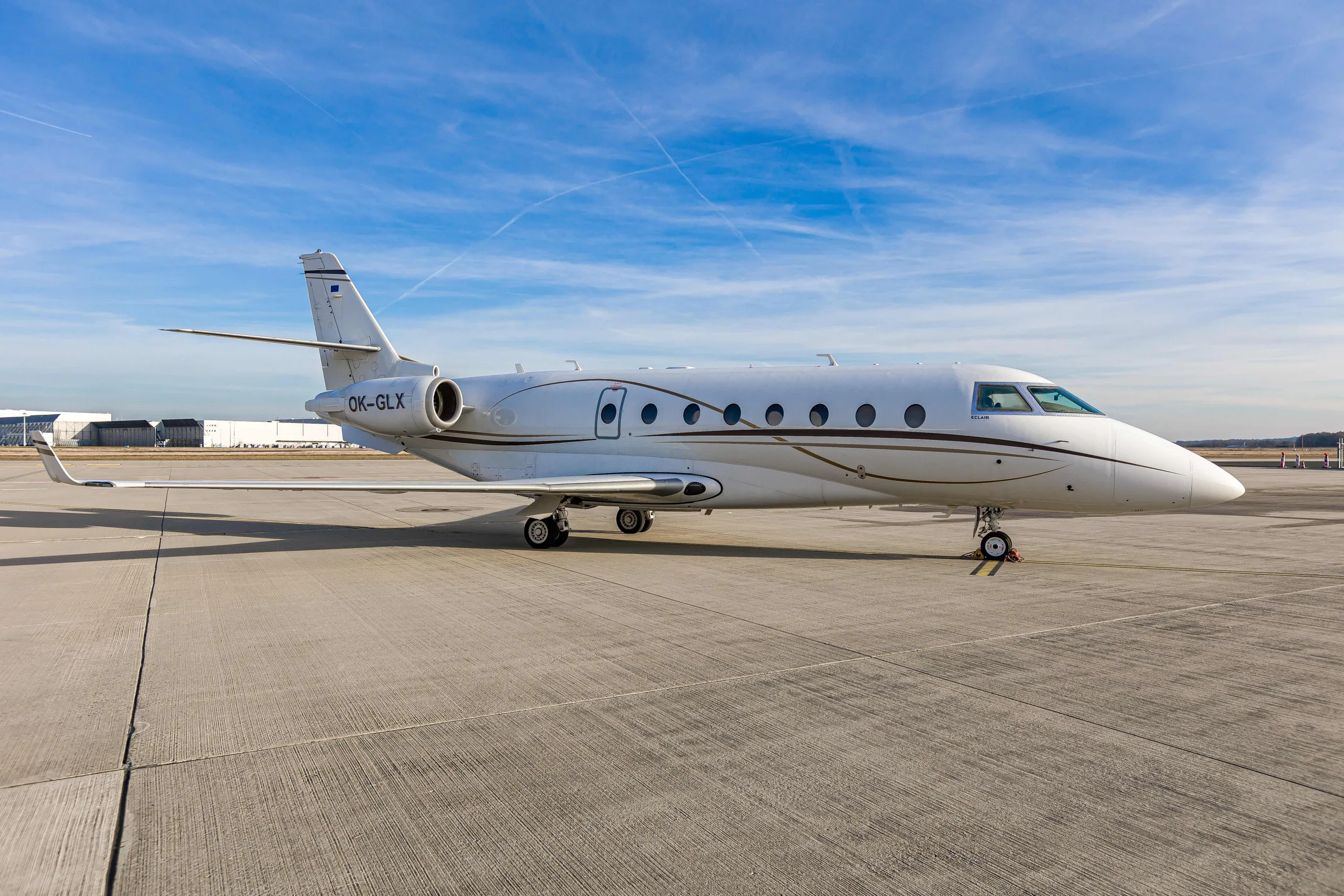 A white Gulfstream G200 business jet with registration OK-GLX parked on an airport tarmac under a clear blue sky.