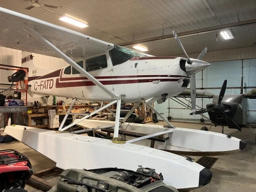 A white floatplane with maroon stripes, registration C-FATD, is parked inside a hangar with another aircraft in the background.