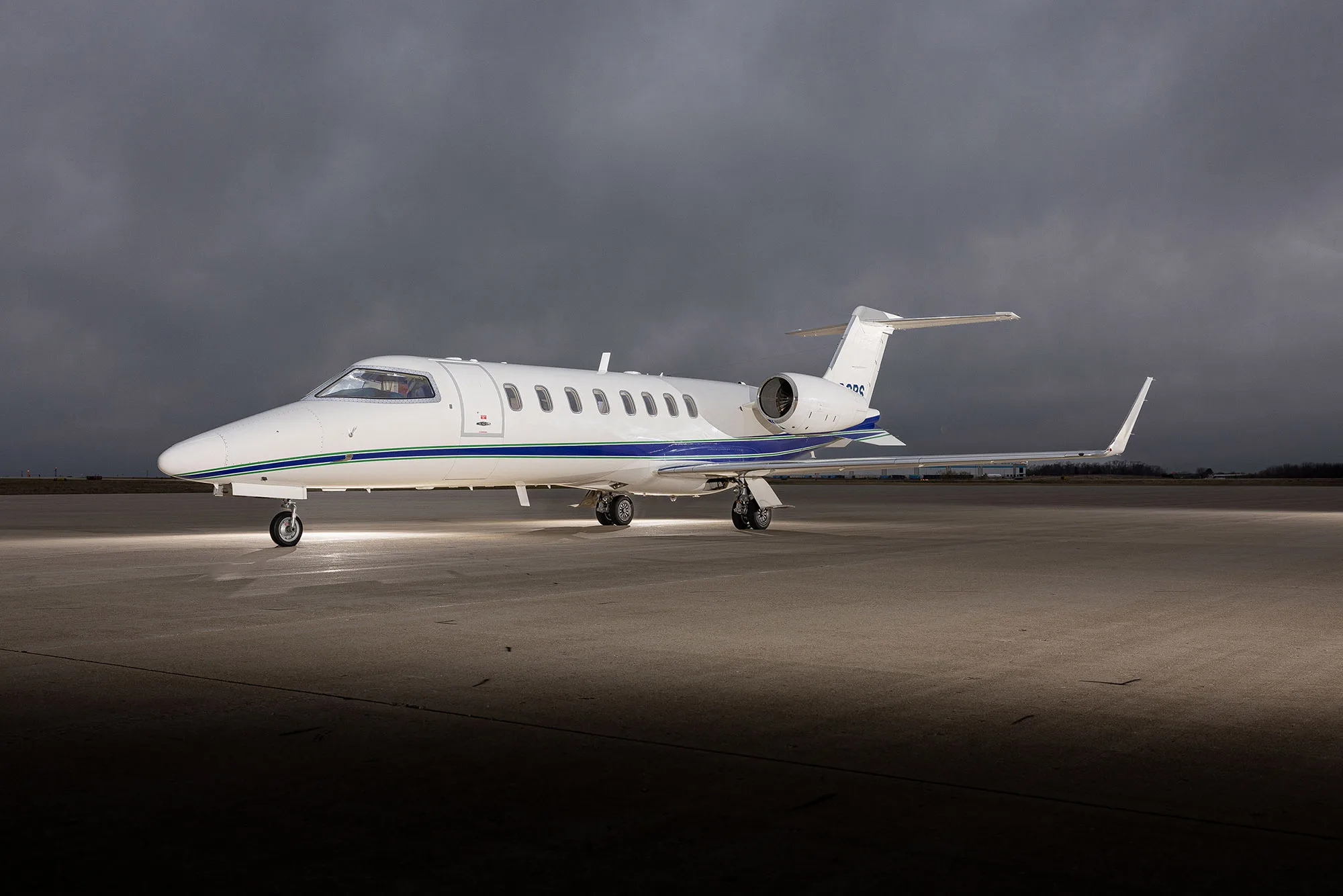 A white Learjet 75 with blue and green stripes parked on an tarmac under dramatic cloudy skies, viewed from the front-left.