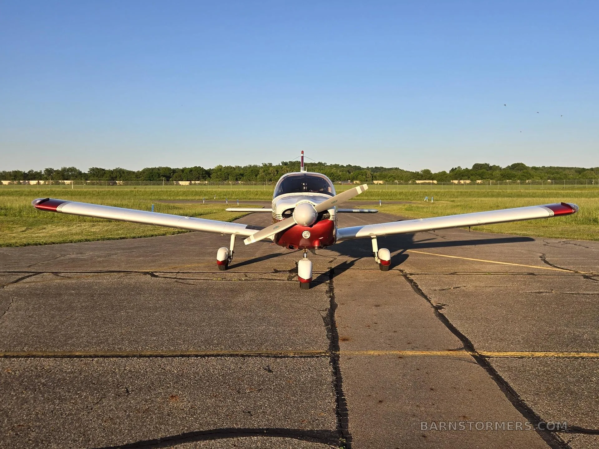 A red and white single-engine propeller aircraft (N55127) is viewed head-on, parked on an asphalt ramp with green grass in the background.
