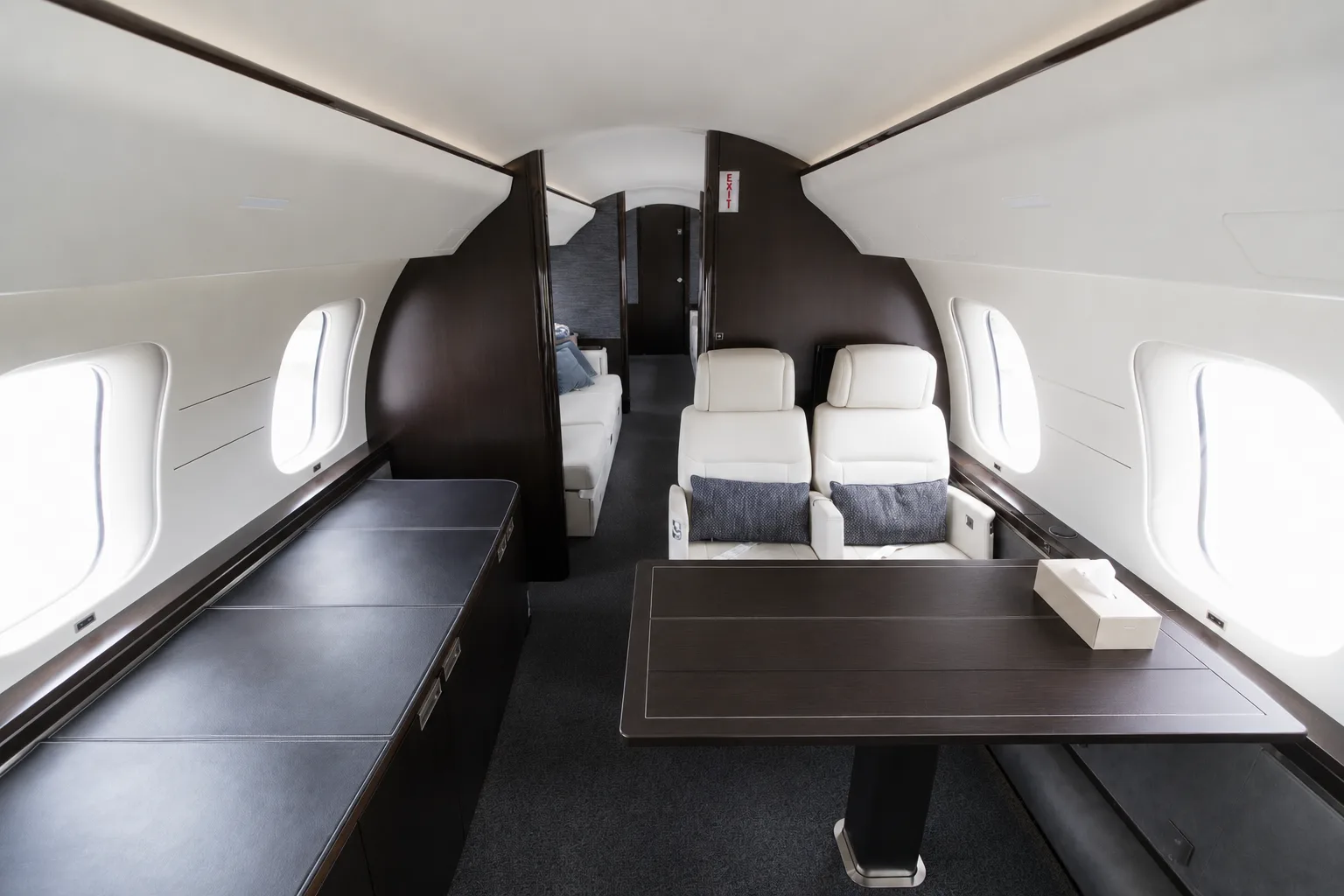 A section of a private jet cabin with a dark wood table, two white leather seats, and a view towards the rear.
