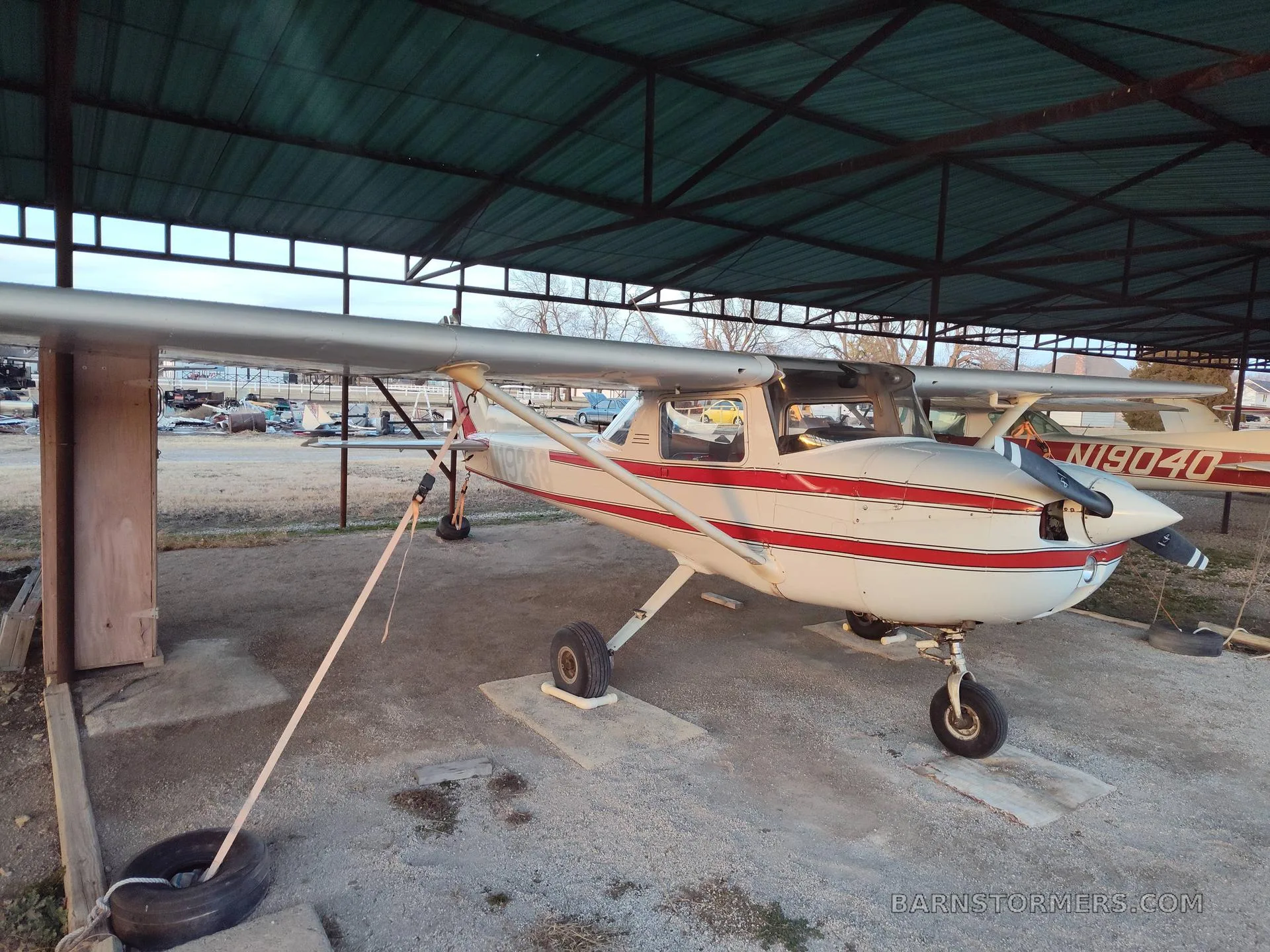 A white and red striped Cessna 150/152 type aircraft is parked under a metal hangar roof, with another aircraft visible in the background.