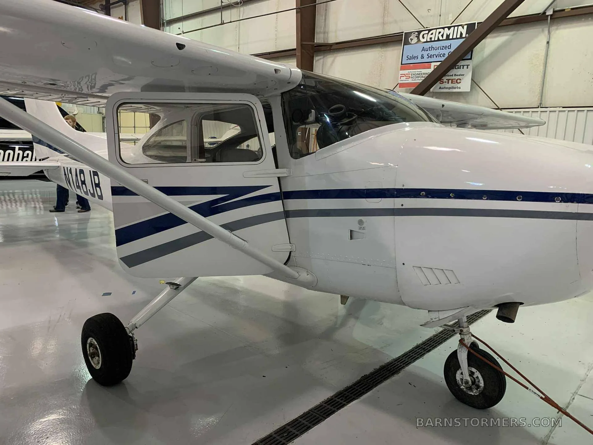 A close-up side view of a white single-engine aircraft with blue and grey stripes, registration N148JB, with its door open in a hangar.