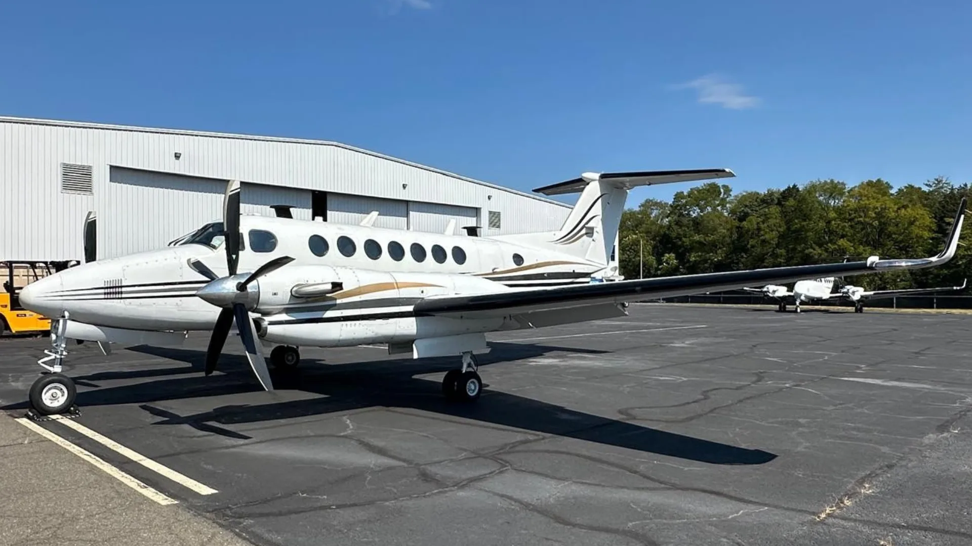 A white Beechcraft King Air with black and gold stripes parked on an asphalt tarmac in front of a hangar.