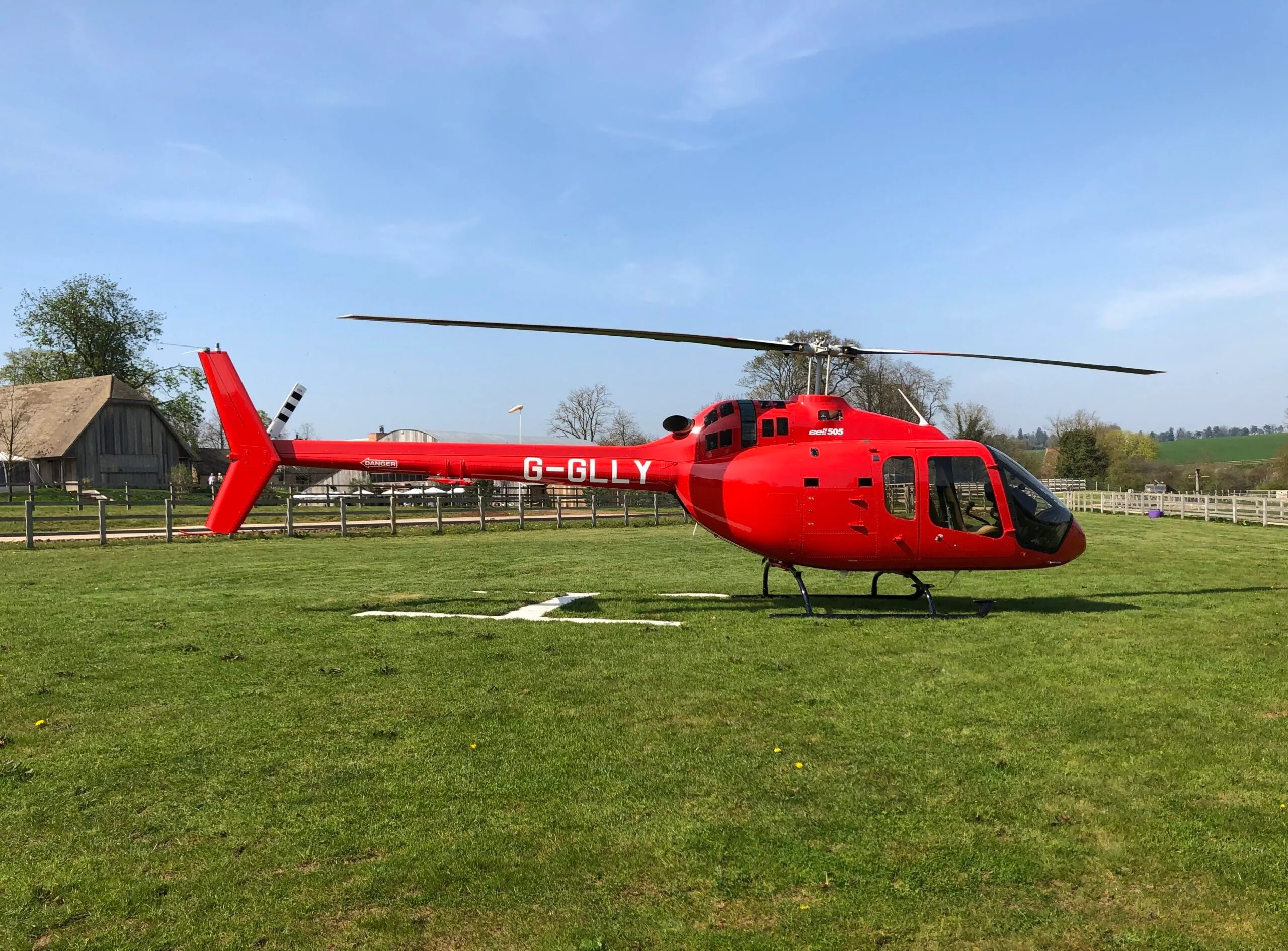 A red Bell 505 helicopter, registration G-GLLY, rests on a green field under a clear blue sky.