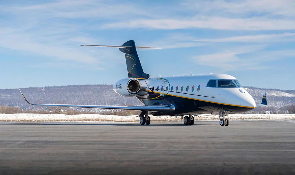 A Gulfstream G550 aircraft, registration N38GL, with a white and dark blue paint scheme, is parked on an airfield with snow-covered hills in the background.