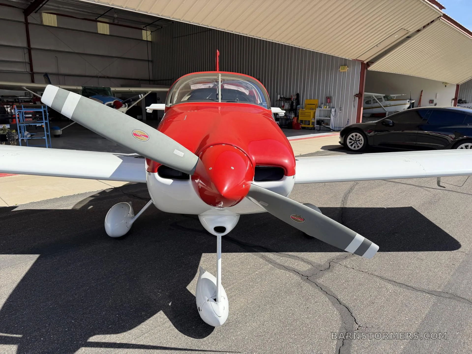 A red and white Vans RV-7A aircraft is parked on the tarmac in front of an open hangar, viewed from the front.