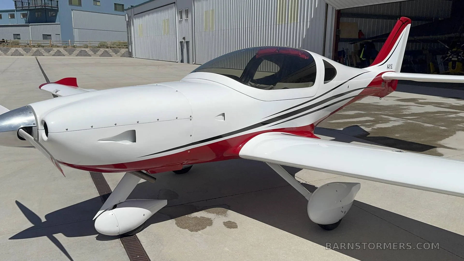 A white and red single-engine aircraft with a bubble canopy, parked on an apron in front of a hangar.