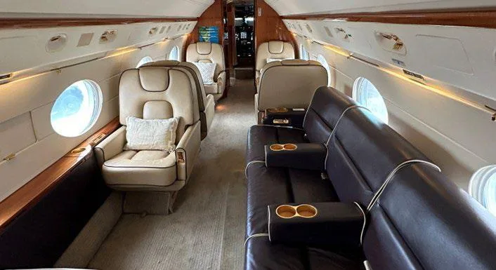 The spacious interior cabin of a business jet featuring a dark leather sofa and individual beige leather seats.