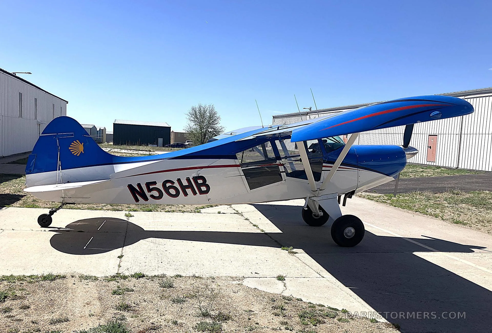A blue and white high-wing aircraft with red stripes and tail logo, registration N56HB, parked on an asphalt apron under a clear blue sky.