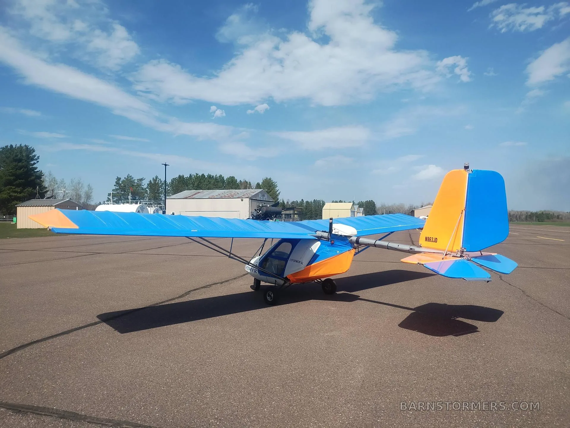 A blue and orange Flightstar II SC ultralight aircraft, tail number N663JD, parked on an airfield under a partly cloudy sky.
