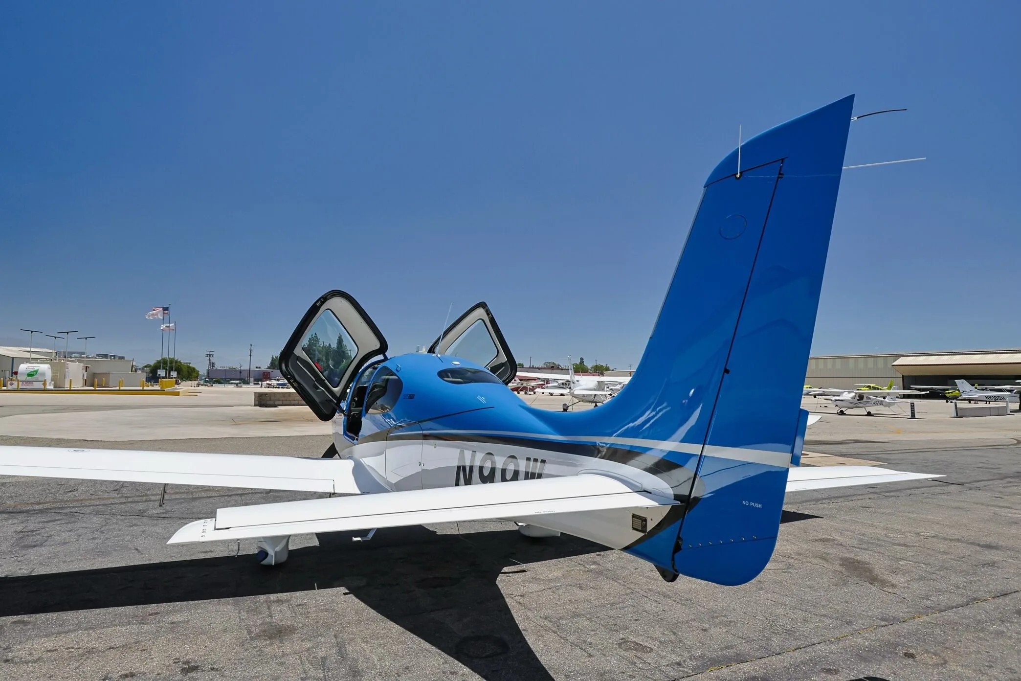 Rear three-quarter view of a blue and white Cirrus SR22 aircraft (N99W) with open doors, parked on an airfield.