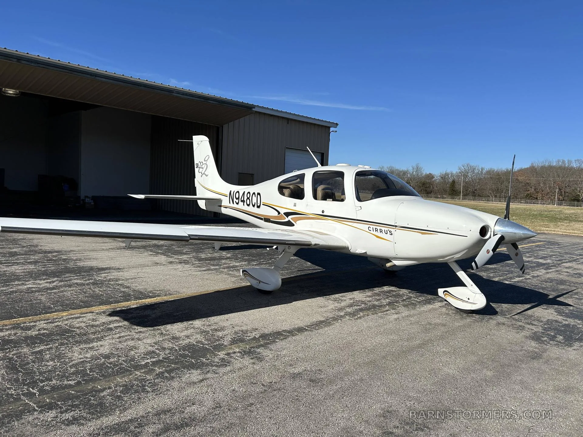 A white Cirrus SR22 aircraft with gold and black stripes, registration N948CD, parked on an asphalt ramp next to a hangar.