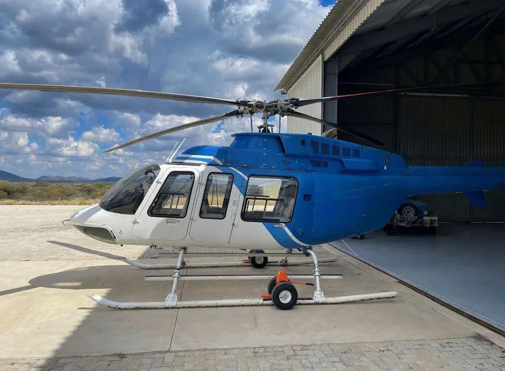 A blue and white helicopter, possibly a Bell 206, parked partially inside a hangar on a sunny day.