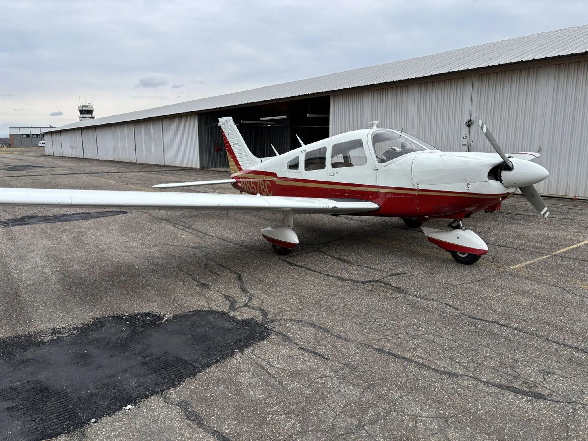 A Piper aircraft, N8578C, with red and gold stripes over white, parked on an airfield with a control tower in the distance.